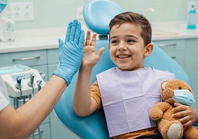 Little boy high-fiving dentist while holding teddy bear.