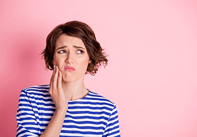 Woman in striped shirt holding hand to jaw in pain with pink background.